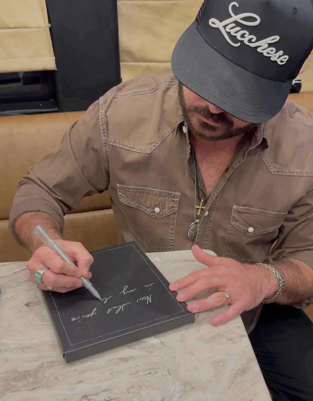Man signing a book with a pen, wearing a 'Lucchese' cap.